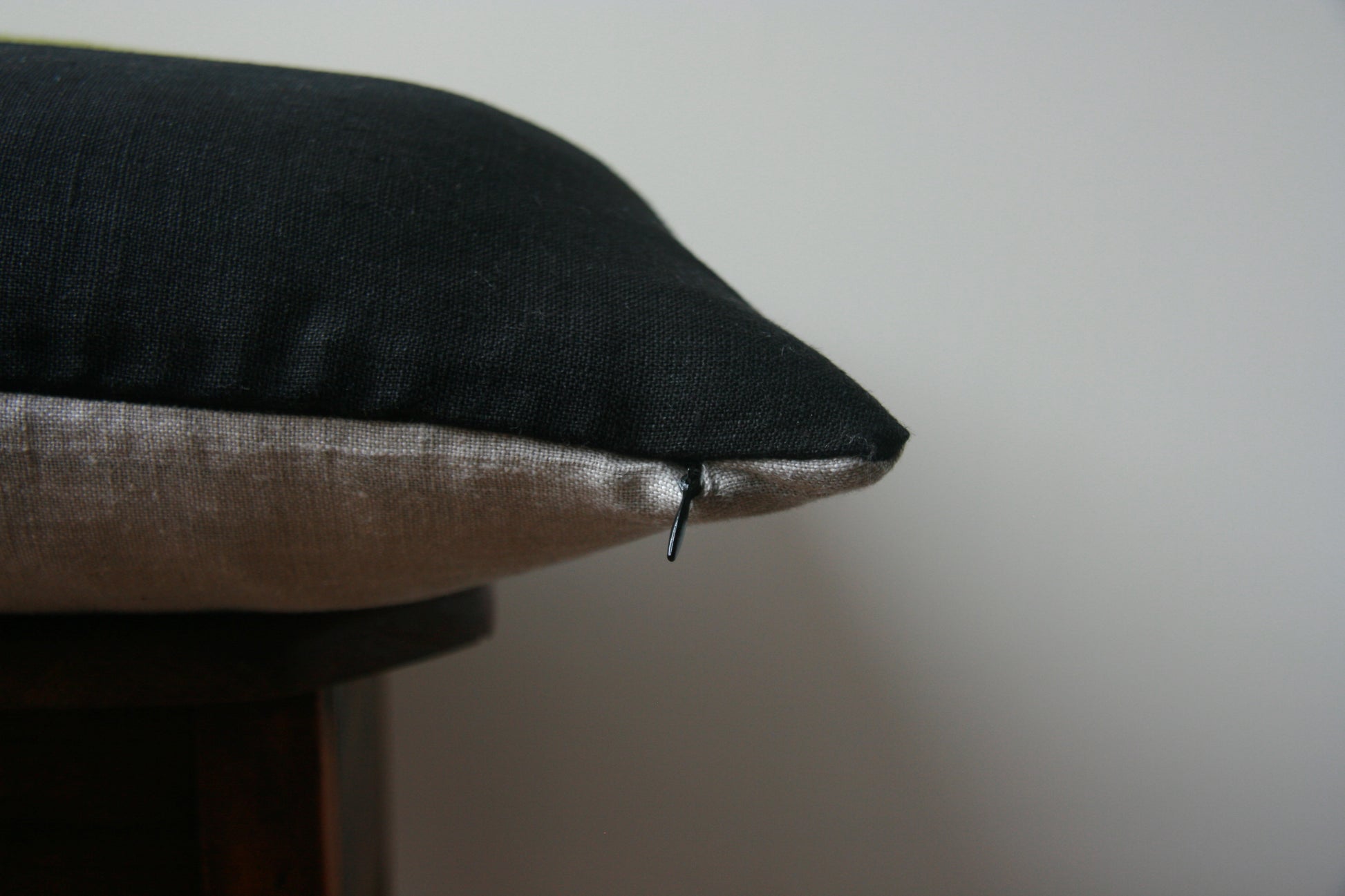 Side profile of cushion cover. Black on top side, natural linen colour on bottom side. Concealed zip with black zipper pull just visible.